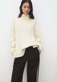 Pull à col roulé couleur crème avec une texture côtelée, manches longues et coupe ample, associé à un pantalon noir avec des bandes latérales.