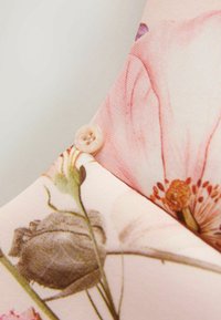 Lähikuva kukkakuvioisesta kankaasta, jonka vaalealle taustalle on ommeltu pieni beige nappi lähelle reunaa.
