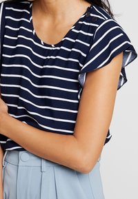 Top blu navy a maniche corte con strisce orizzontali bianche, maniche arricciate e due bottoni sulla spalla. Tessuto morbido e leggero.