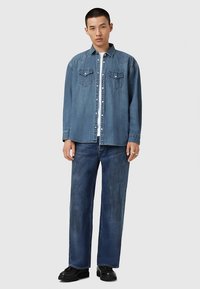 Giovane uomo che indossa una camicia di jeans blu sopra una maglietta bianca, abbinata a jeans dritti blu scuro e scarpe nere, in piedi contro uno sfondo semplice.