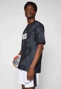 Sort og grå t-shirt med korte ærmer, der har et abstrakt mønster og teksten "PROD/CER", kombineret med hvide shorts. Personen holder en tekstureret fodbold.