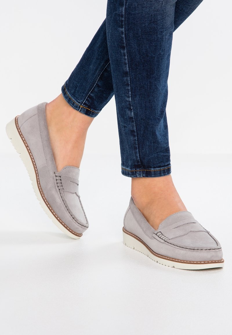 Pieds portant des mocassins en daim gris clair avec des semelles blanches et un jean bleu foncé retroussé sur un fond blanc.