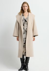 Beige uldfrakke med brede ærmer, store revers og synlige sømdetaljer. Underfrakken en mønstret kjole i sort, hvid og beige nuancer.