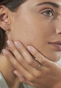 Boucles d'oreilles en or avec de petits charms ; une bague en or texturée sur une main. Peau lisse et finition naturelle des ongles. Arrière-plan flou.