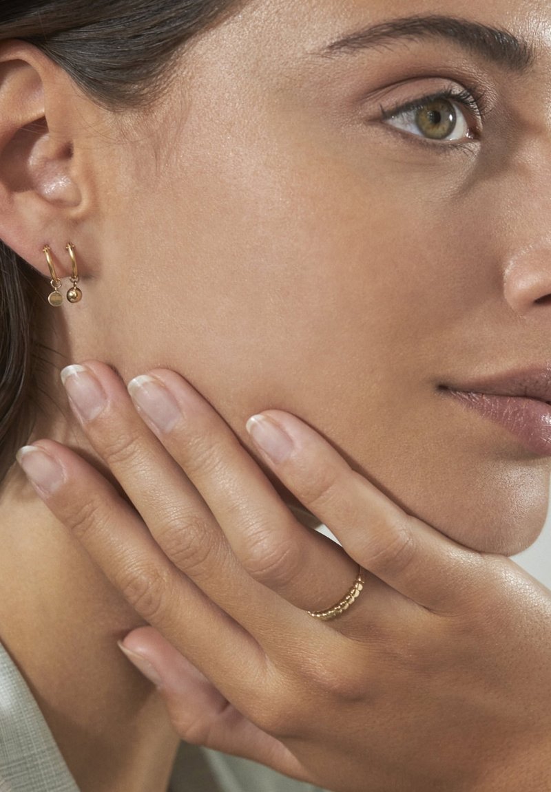 Gouden creolen met kleine bedeltjes; een getextureerde gouden ring op een hand. Gladde huid en een natuurlijke nagelfinish. Zachte achtergrond in vervaging.