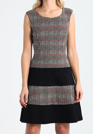 Mouwloze jurk tot op de knie met een zwart-rood geruit patroon op de bovenkant en het midden, effen zwarte horizontale banden, gedragen door een staande vrouw.