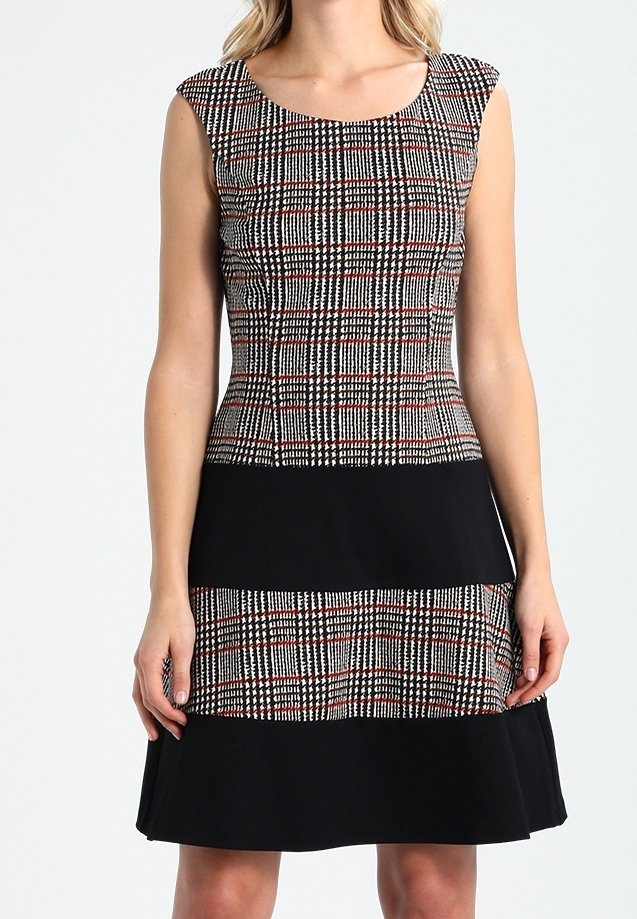 Robe sans manches longueur genou avec motif à carreaux noir et rouge sur le haut et la partie médiane, bandes horizontales noires unies, portée par une femme debout.