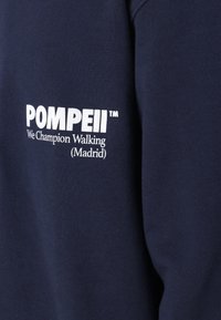 Marinegenser med hvit trykt tekst "POMPEII™ We Champion Walking (Madrid)" på fronten. Myk tekstur, rund halsdesign og lange ermer.