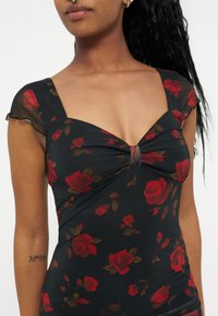 Femme portant une robe noire avec des motifs de roses rouges, dotée d'un buste froncé et de manches courtes transparentes.