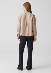 Beige overhemd met lange mouwen, een split aan de achterkant, een kraag en knopenmanchetten, gecombineerd met zwarte op maat gemaakte broek. Gladde textuur, neutrale kleuren.