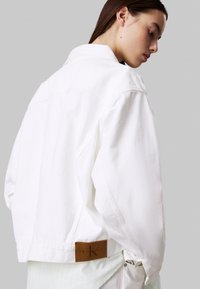 La chaqueta de mezclilla blanca presenta un corte relajado, cuello puntiagudo y una etiqueta de cuero marrón de Calvin Klein Jeans en la parte de atrás. Textura suave.