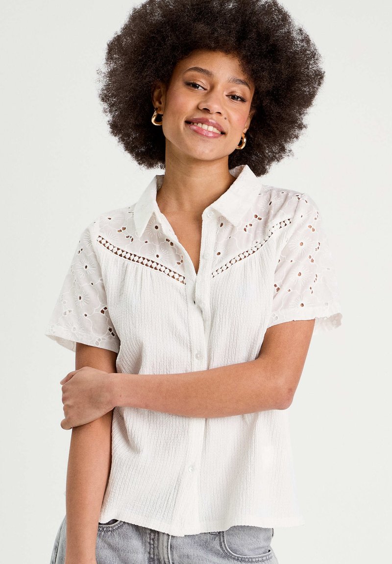 Chemise blanche à manches courtes avec un corps texturé et des manches en dentelle à œillets brodés. Présente un devant à boutons et une coupe décontractée.