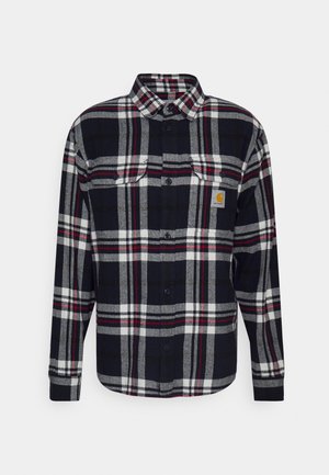Chemise à carreaux noire, blanche et rouge à manches longues, boutonnée, avec deux poches avant et un petit patch logo jaune sur la poche poitrine gauche.