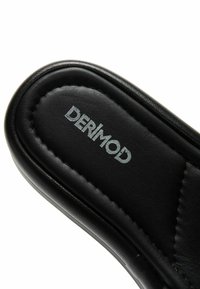Gros plan sur la semelle rembourrée noire d'une pantoufle avec le nom de la marque « DERIMOD » imprimé en argent sur la zone du talon.