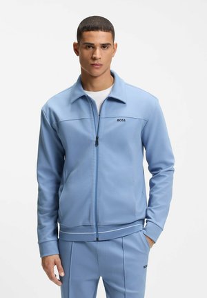 Hombre con chaqueta azul claro con cremallera y pantalones a juego con el logo "BOSS", de pie con una mano en el bolsillo contra un fondo blanco.