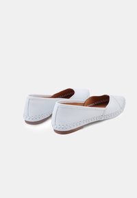 Zapatos slip-on de cuero blanco con punta redondeada, costuras decorativas a lo largo de los bordes y una suela plana de goma marrón.