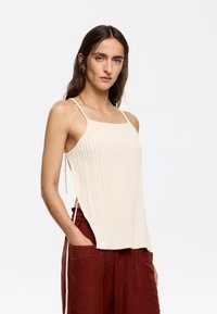Light beige knitted tank top with thin straps, ribbed texture, and side slits. Paired with high-waisted rust-colored trousers.