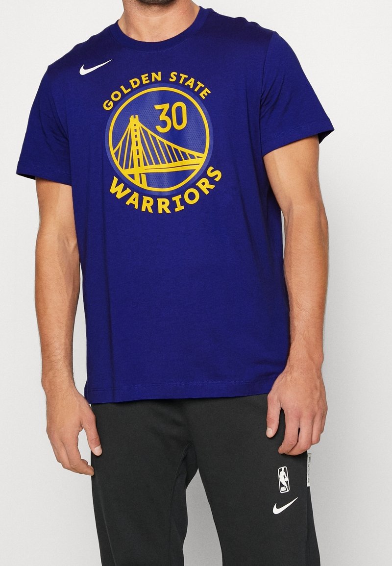 Homme portant un t-shirt bleu des Golden State Warriors avec le numéro 30 et un pantalon de survêtement noir NBA Nike, debout devant un fond clair uni.