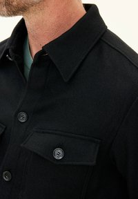 Black button-up shirt with a collar, featuring a textured fabric, two chest pockets, and branded buttons.