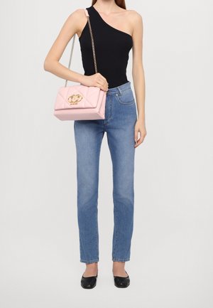 Mujer con top negro de un solo hombro, jeans azules, zapatos planos negros, sosteniendo un bolso rosa acolchado con cadena dorada y cierre con logo.