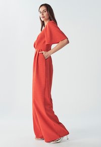 Mujer con un mono rojo fluido, de piernas anchas y mangas fluyentes, de pie de lado con una mano en el bolsillo, contra un fondo liso.