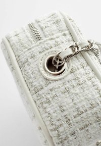 Witte geweven handtas met metalen draden, voorzien van een zilveren rits, een ronde goudkleurige ring en een zilveren schouderband. Getextureerd oppervlak.