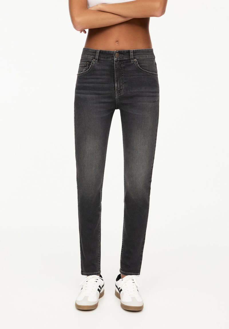 Jeans neri skinny con un design a vita media, dotati di tasche anteriori e cuciture tonali, abbinati a sneakers bianche con rifiniture nere.
