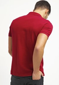 Chemise polo rouge à manches courtes, texture lisse, et col classique, portée à l'envers, montrant un design uni sans logos ni motifs.