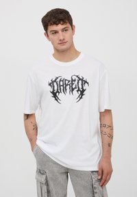 Biała bawełniana koszulka z krótkim rękawem, z czarnym graficznym logo na środku. Model nosi jasnoszare spodnie cargo.