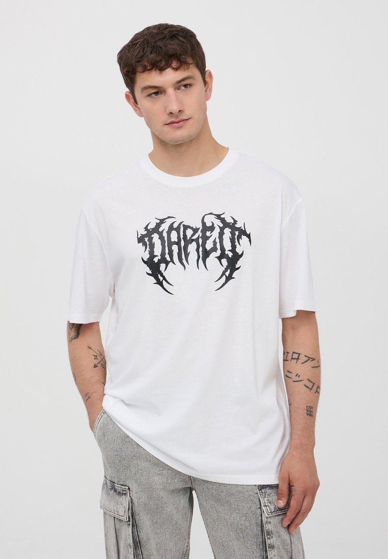 Biała bawełniana koszulka z krótkim rękawem, z czarnym graficznym logo na środku. Model nosi jasnoszare spodnie cargo.