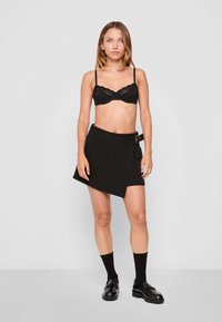 Black lace bra with adjustable straps paired with a black wrap skirt featuring a side buckle and flat hem. Black ankle socks and loafers complete the look.