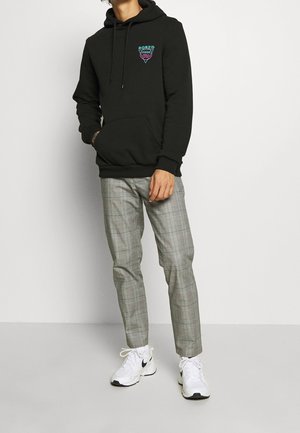 Personne portant un sweat à capuche noir avec le texte "Bored Inside", un pantalon à carreaux gris et des baskets blanches Nike, debout devant un fond blanc.