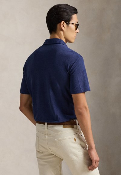 Πόλο μπλούζα ναυτικού μπλε με κοντά μανίκια, υφασμάτινη υφή και άνετη εφαρμογή, συνδυασμένη με ανοιχτόχρωμο παντελόνι και καφέ ζώνη.