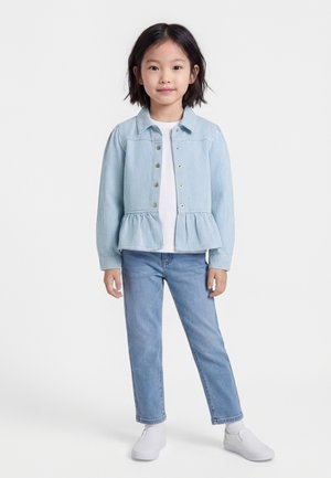 Νεαρή κοπέλα όρθια, φοράει ανοιχτό μπλε ριγέ μπουφάν, λευκό πουκάμισο, μπλε τζιν και λευκά παπούτσια τύπου slip-on, με φόντο μια απλή επιφάνεια.