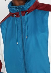 Veste coupe-vent avec une fermeture éclair à l'avant, un col montant et un design en blocs de couleur en teal, bordeaux et blanc, avec des cordons ajustables.