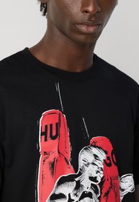 Prim-plan cu o persoană îmbrăcată într-un tricou negru, având un grafic cu un pugilist care lovește mănuși de box roșii, alături de textul "HUGO".