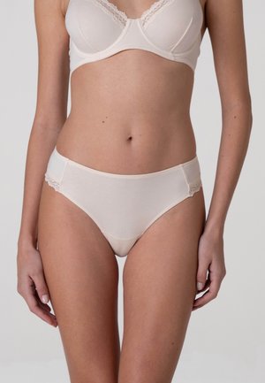 Donna che indossa un reggiseno beige con bordo in pizzo e mutandine abbinate, in piedi su uno sfondo chiaro e uniforme.