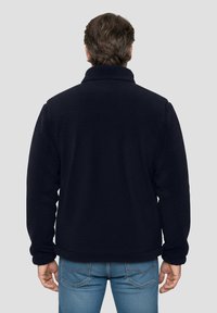 Marineblaue Fleecejacke mit hohem Kragen und strukturierter Stoffqualität. Verfügt über eine lockere Passform, keine sichtbaren Verschlüsse und wird mit hellblauen Jeans kombiniert.