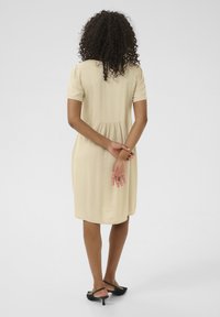 Abito beige a maniche corte con corpetto aderente e orlo svasato, realizzato in un tessuto morbido. Tacchi neri completano l'outfit.