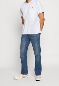 Hombre con camiseta de manga corta a rayas azul claro y blanca, jeans azules y zapatillas blancas, de pie con las manos en los bolsillos.