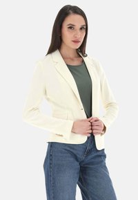 Blazer crema con vestibilità tailored, chiusura a singolo bottone e due tasche laterali, abbinato a una maglietta verde e jeans blu a vita alta.