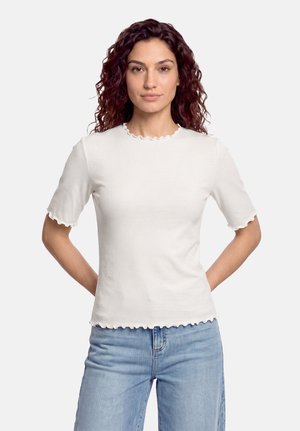 Kvinde med krøllet mørkt hår iført en hvid kortærmet skjorte med flæsekant og lyseblå jeans, står med hænderne bag ryggen.