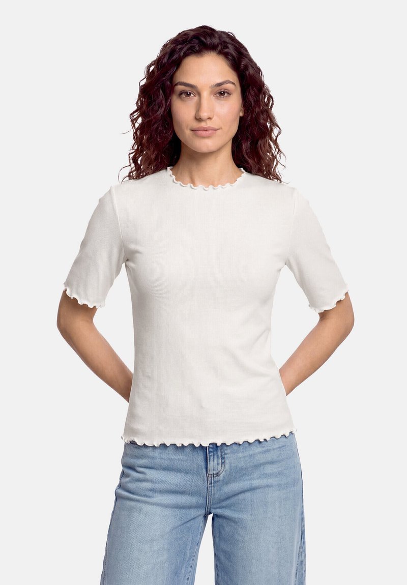 Femme aux cheveux bouclés et foncés portant une chemise blanche à manches courtes avec des bords volantés et un jean bleu clair, debout les mains dans le dos.
