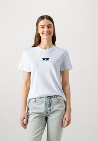 Balta medvilninė marškinėlių su trumpomis rankovėmis ir apvalia iškirpte, su juodu grafiniu dizainu, vaizduojančiu akinius nuo saulės ir žodžiu "KARL LAGERFELD" po jais.