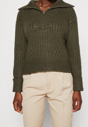 Pull en maille côtelée vert foncé avec col à demi-fermeture éclair et manches longues, associé à un pantalon beige clair.