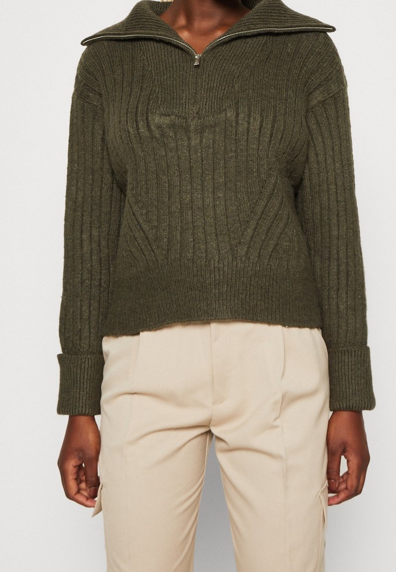 Pull en maille côtelée vert foncé avec col à demi-fermeture éclair et manches longues, associé à un pantalon beige clair.
