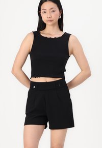Top corto negro con escote en pico ondulado combinado con pantalones cortos negros de tiro alto que presentan dos botones delanteros y una textura suave.