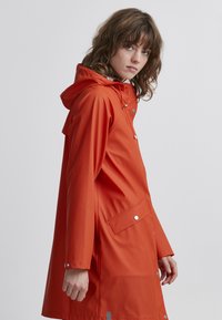 Impermeabile arancione con cappuccio, bottoni a scatto e tasca frontale. Tessuto liscio con un design leggermente aderente.