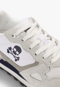 Zapatilla deportiva blanca con acentos de gamuza gris, material sintético texturizado, logo prominente de calavera y huesos cruzados, y cordones blancos.