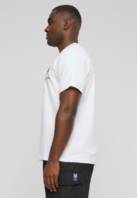 Hvid bomulds t-shirt med korte ærmer, prydet med et sort grafisk print på forsiden. Båret sammen med sorte cargo bukser, vist i profil.